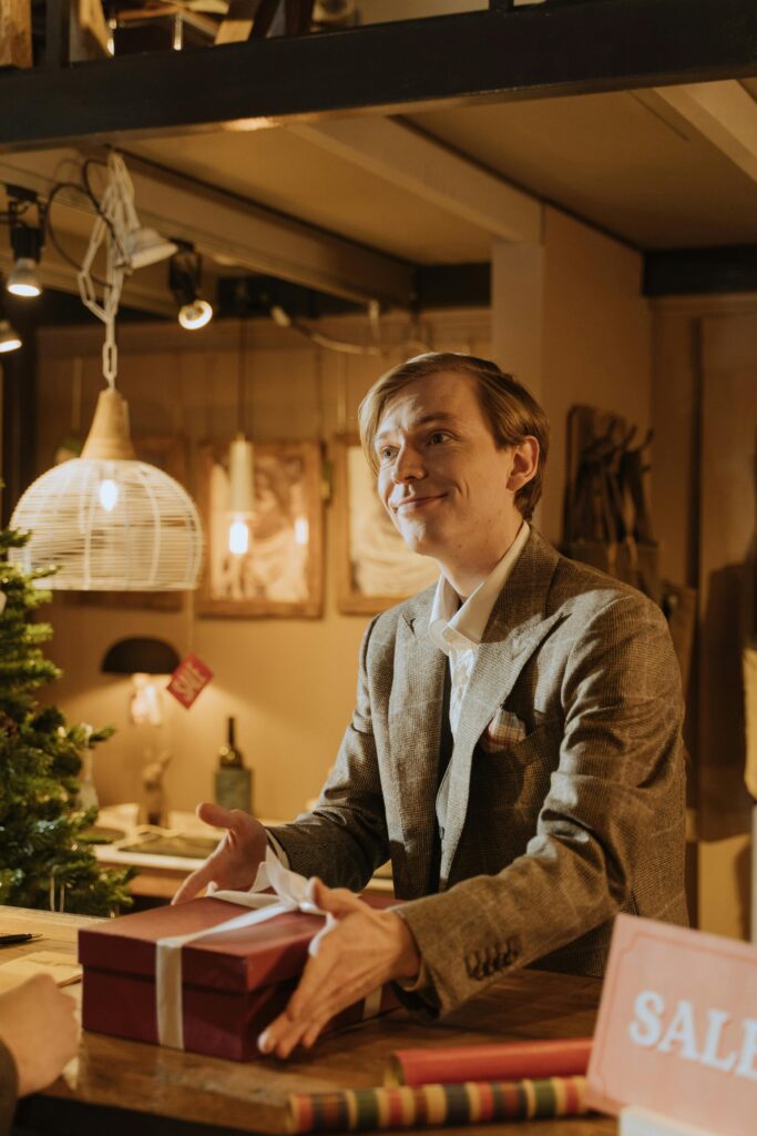 Smiling businessman in suit presenting a wrapped gift in a warmly lit shop with Christmas decor.