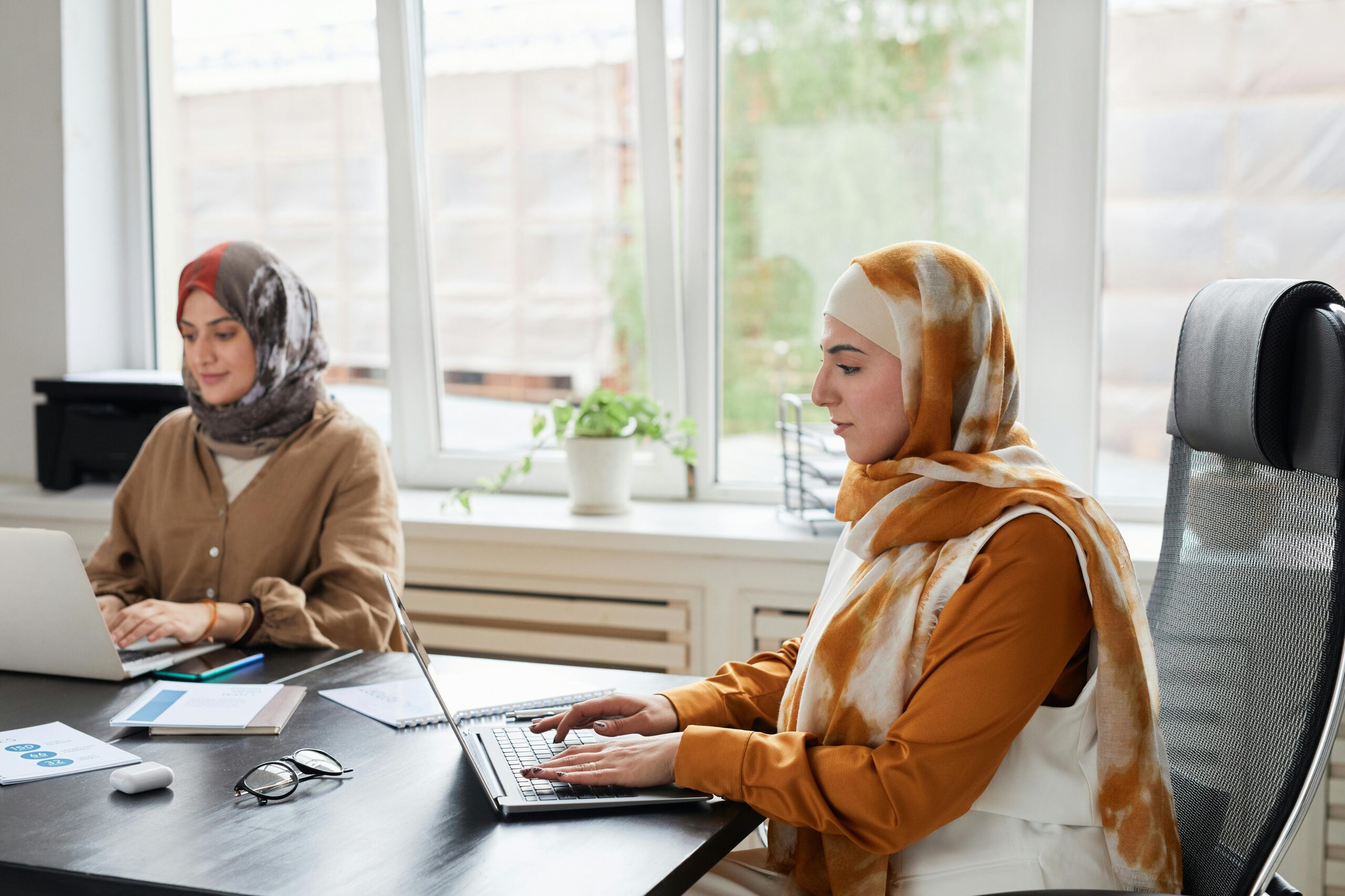 Two Muslim women working on laptops in a modern office setting.